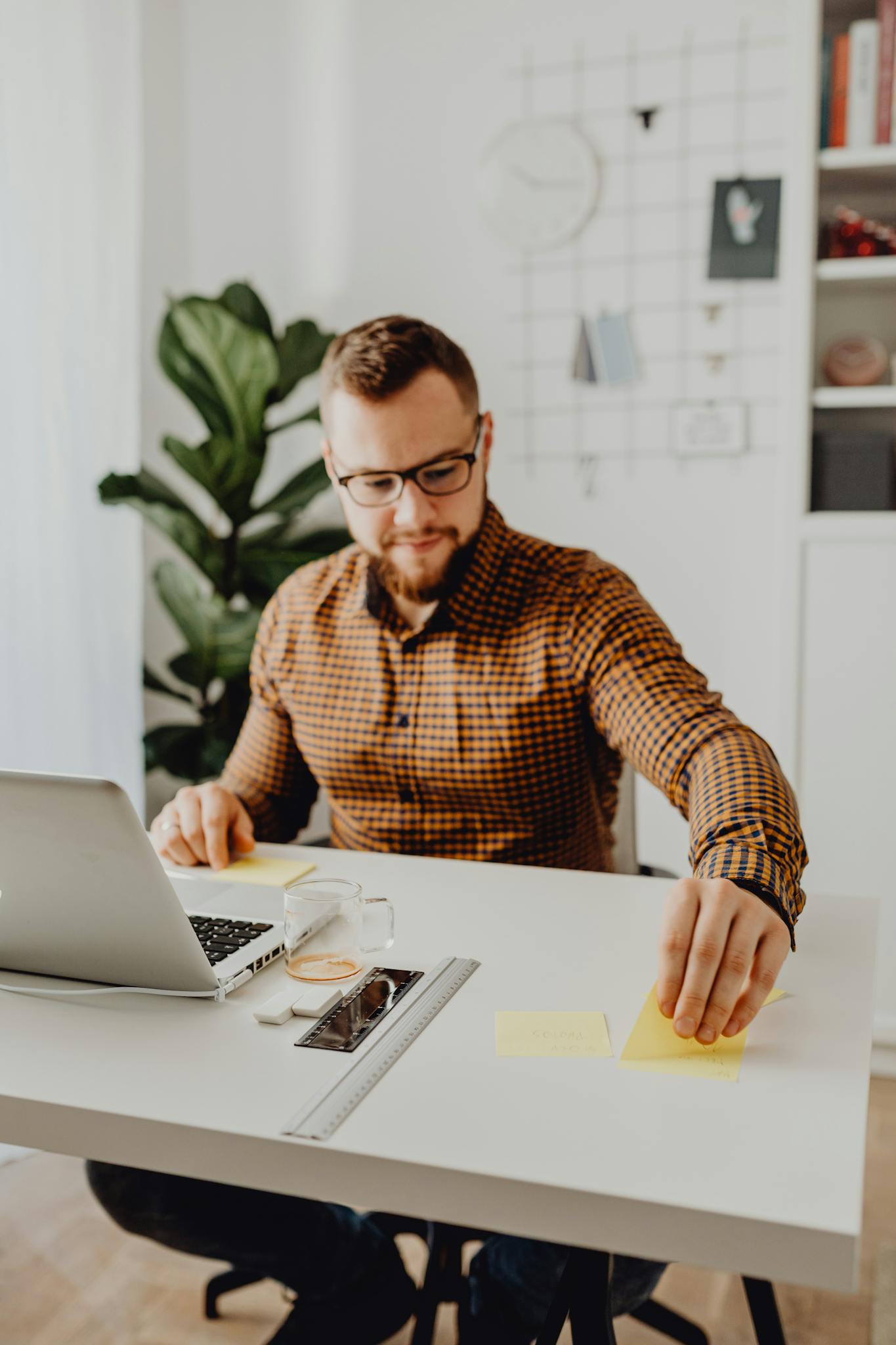 Adult man using sticky notes at his workstation with laptop, eyeglasses, and ruler on desk.