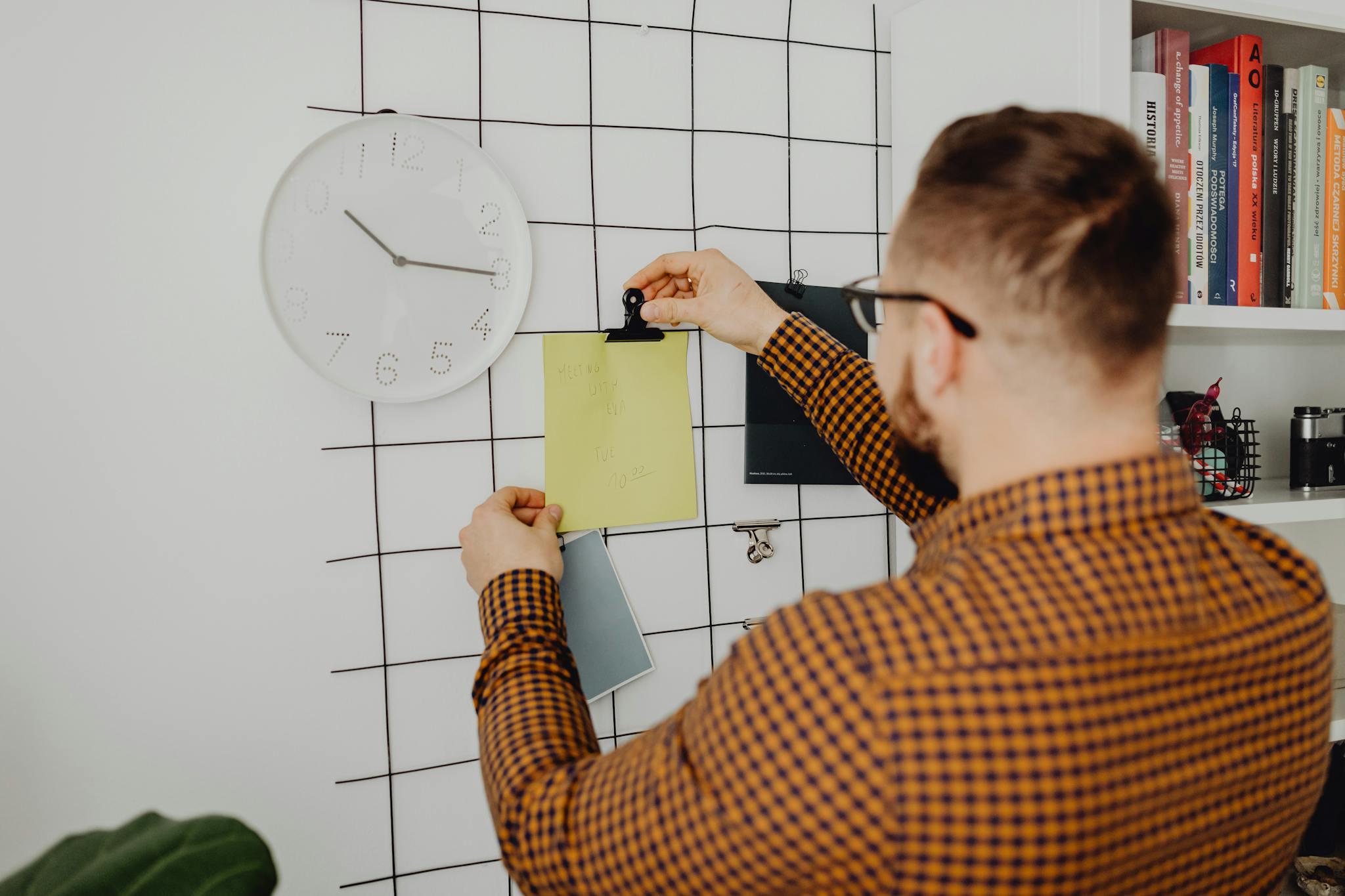 Person arranging notes on an office wall grid beside a clock, creating an organized workspace.